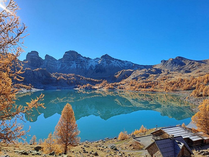 Lake Allos in autumn, with the vegetation around the lake turning brown and the larch trees turning orange, and the mountains known as the Tours du Lac in the background, covered in patchy snow